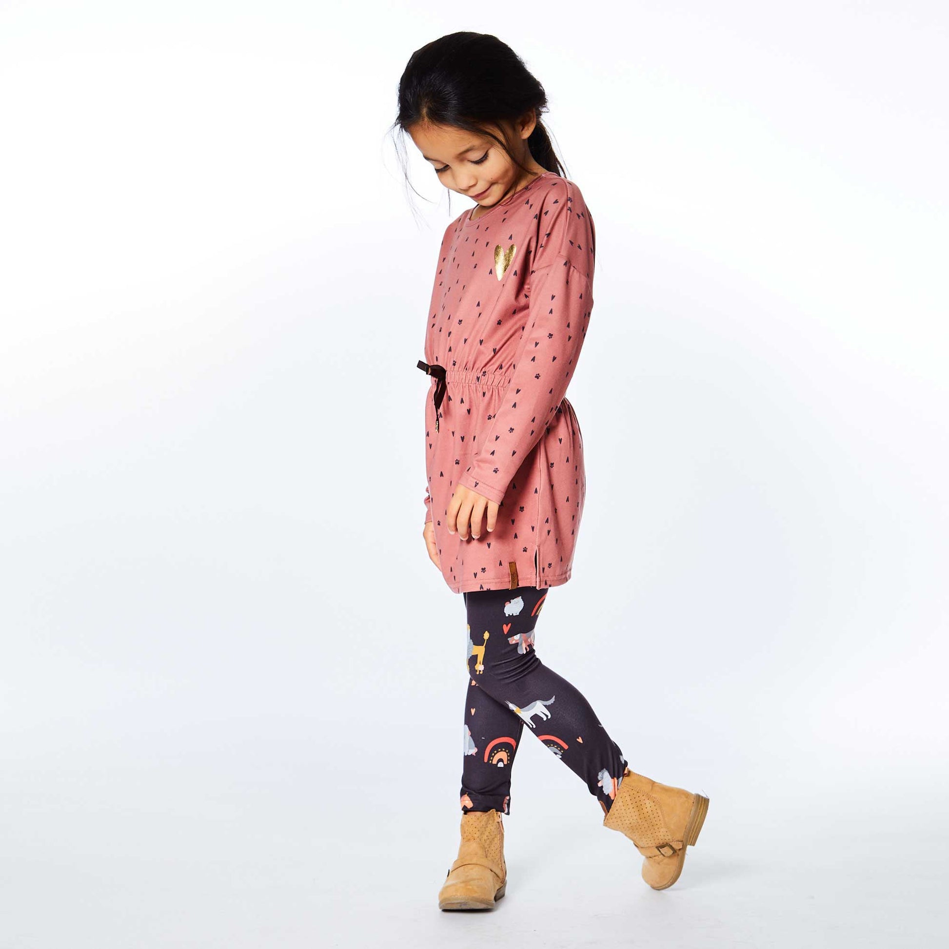 An allover heart print with puppy paws makes our Printed Paws and Hearts Long Sleeve Tunic a fun addition to any little girl's wardrobe.  This adorable top is made from soft stretch poly fabric and features long sleeves with dropped shoulders, a round neckline, and an elastic waistband with a ribbon bow and a dog bone embellishment.